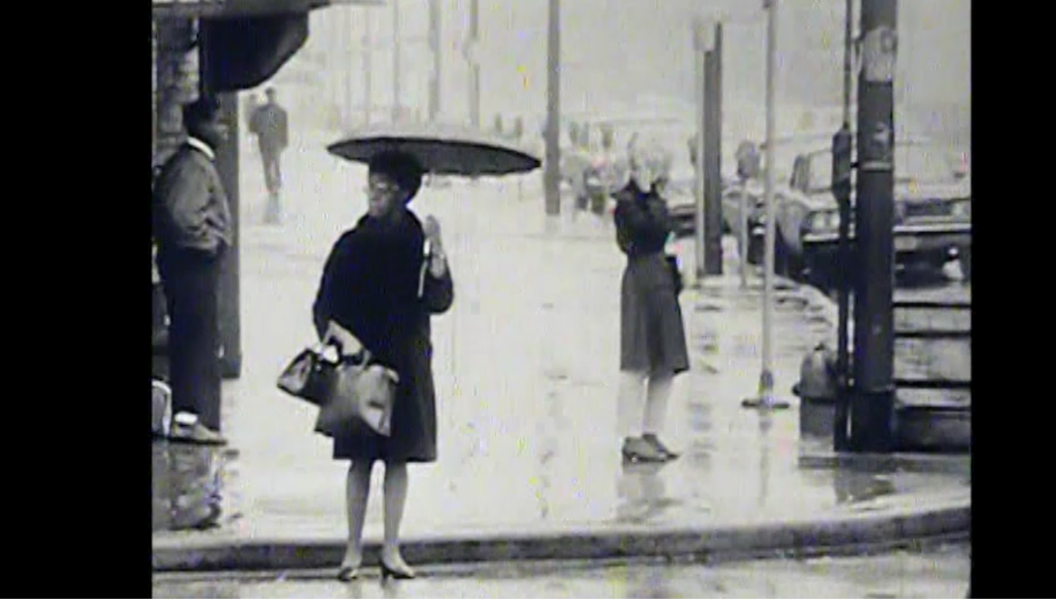 Black-and-white still image of a rainy urban street scene. In the foreground, a woman stands at a corner holding an umbrella and carrying two bags, wearing a dark coat and heeled shoes. She looks to her left while waiting. Behind her across the street, another woman stands on the wet sidewalk near a utility pole, also appearing to wait. The background is filled with blurred pedestrians, utility poles, parked cars, and reflections on the wet pavement, capturing the quiet atmosphere of a rainy day.
