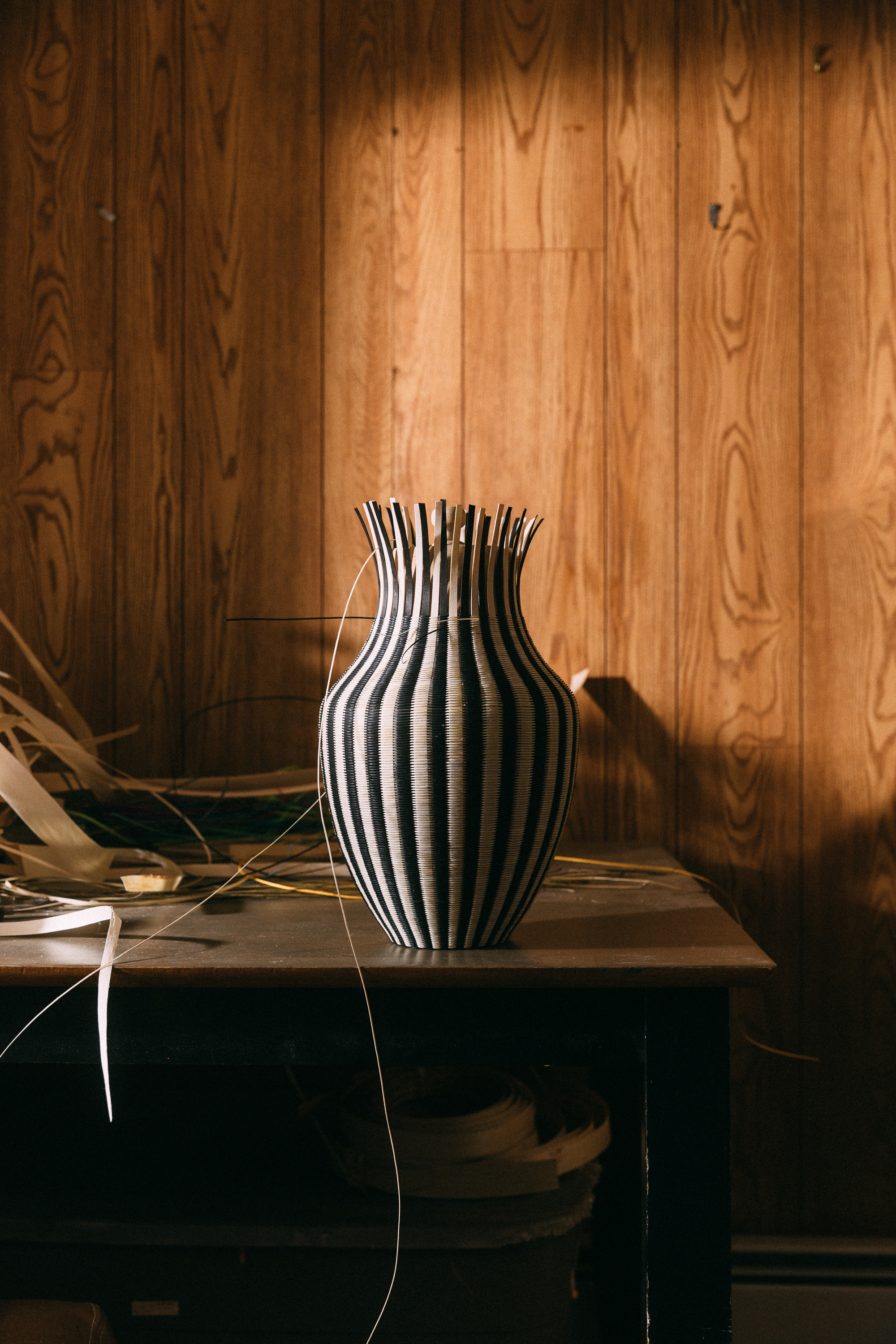 A color photo shows a two-tone tall basket with blond and black vertical stripes. The edges of the vertical strips are not bound at the top. The basket sits on a surface next to strips of wood of varying widths.