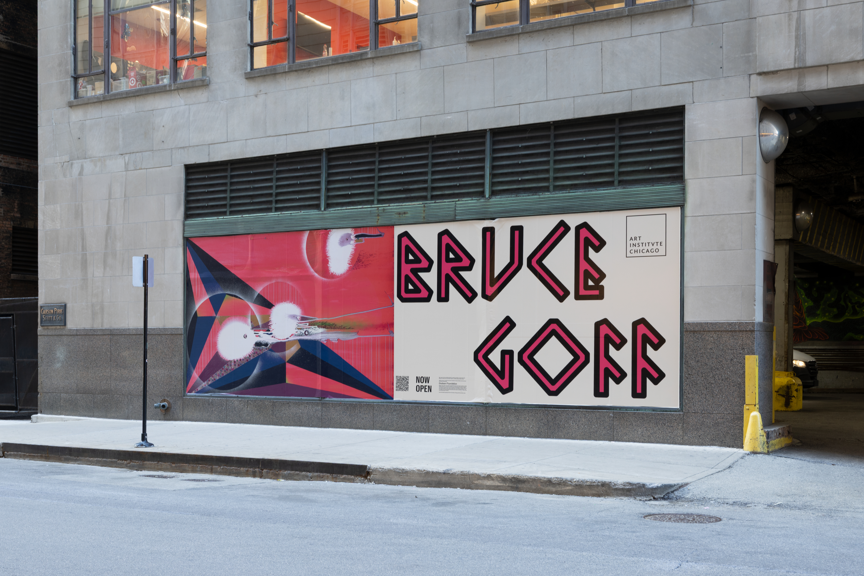 Along the face of a gray building on a city street is a large wall advertisement featuring a red artwork with blue and black geometric shapes and white circles at left and, at right, a cream panel with bold red-and-black writing that reads “BRUCE GOFF.”