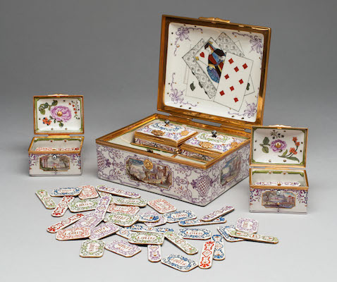 A richly decorated gaming box mounted with gold plaques and painted with violet and gold enamels and gilding. The box is open and reveals four small, similarly decorated porcelain containers. These hold two types of porcelain gambling chips. The lid of the large box depicts three playing cards that appear to have been carelessly thrown down.