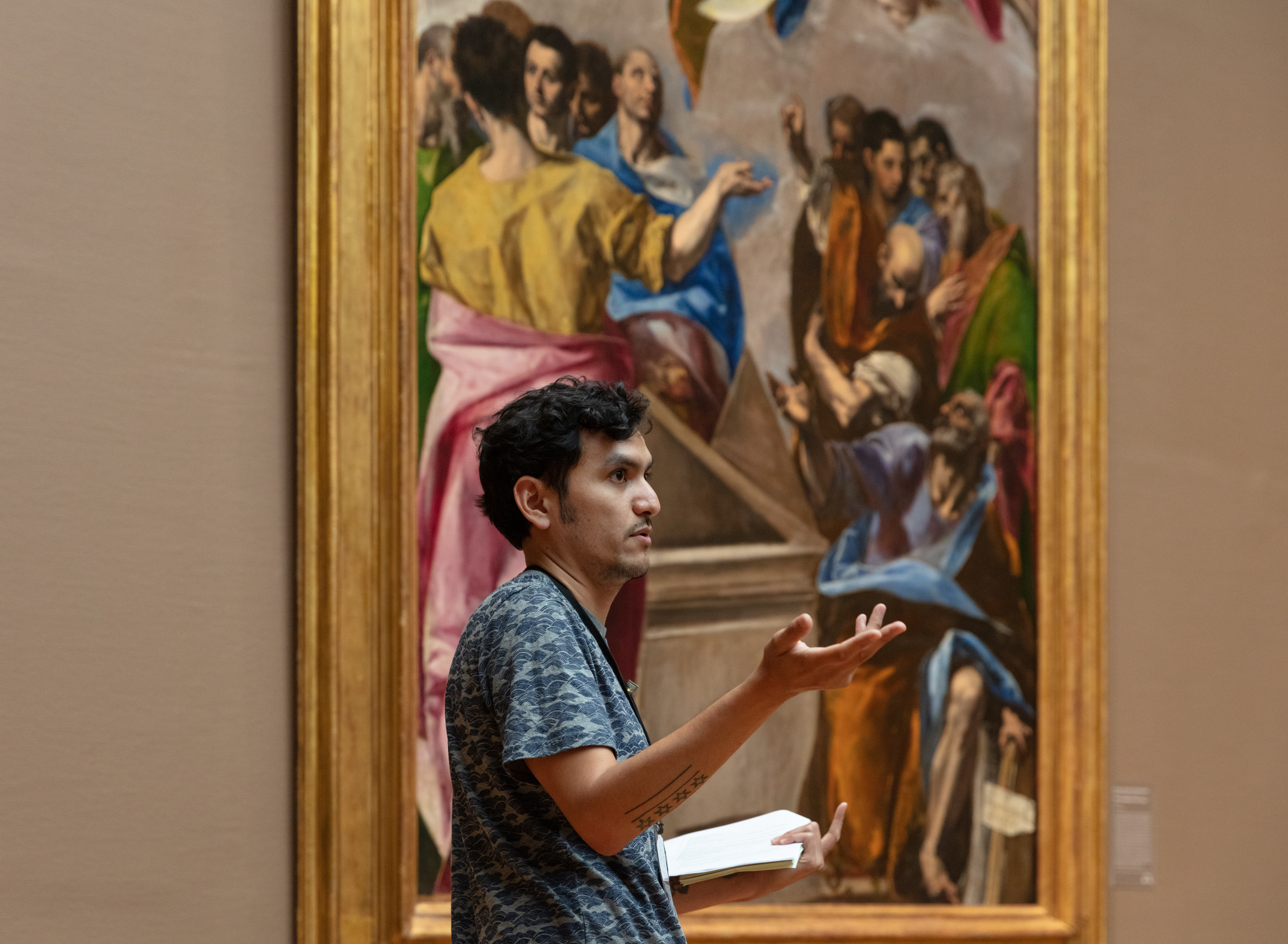 A medium-skinned man with dark hair, Sam Ramos, stands pointing before El Greco’s "The Assumption of the Virgin," an enormous canvas featuring numerous robed figures.