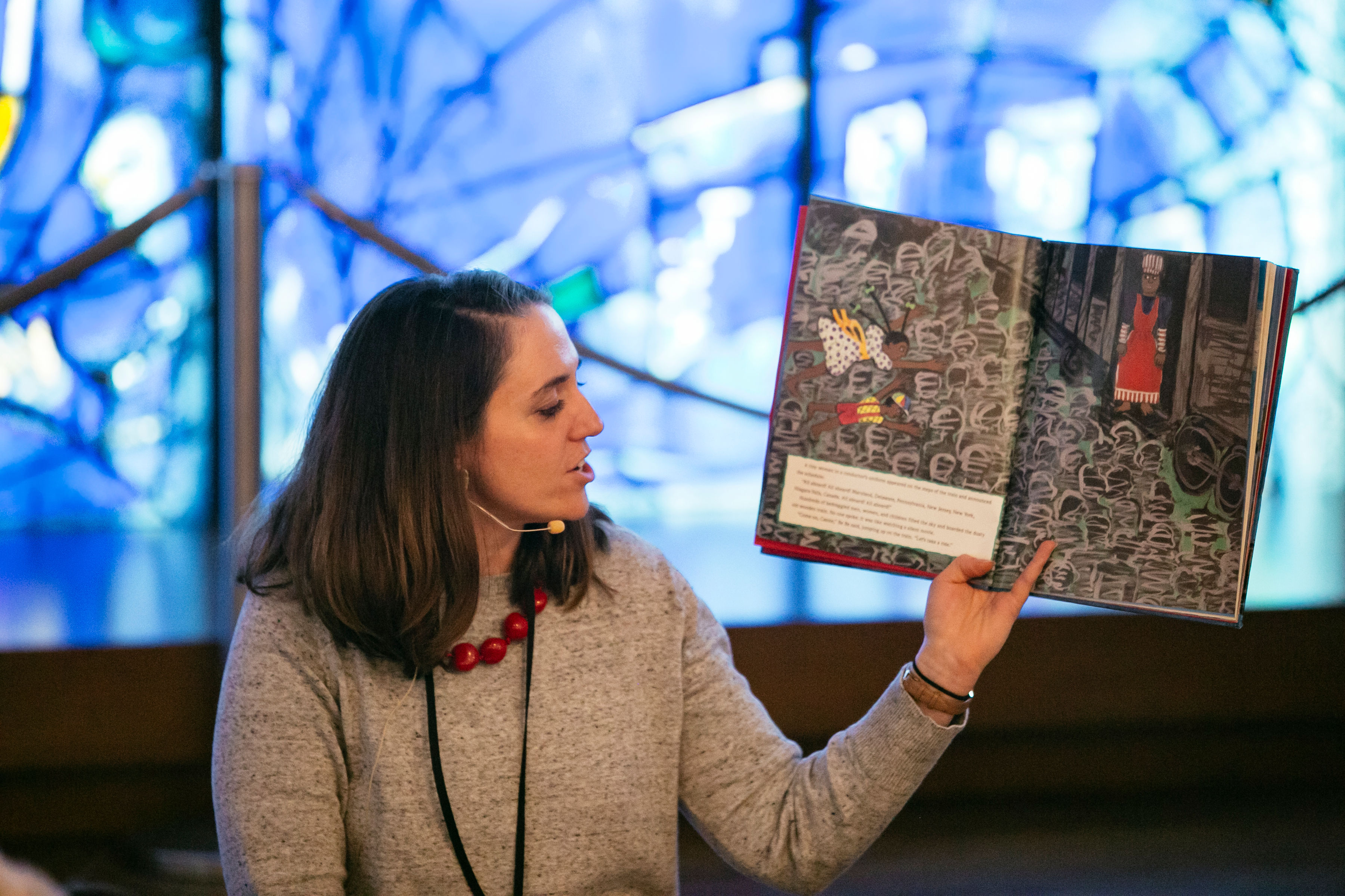 A light-skinned woman holds up a picture book for her audience to see while looking over at it reading.