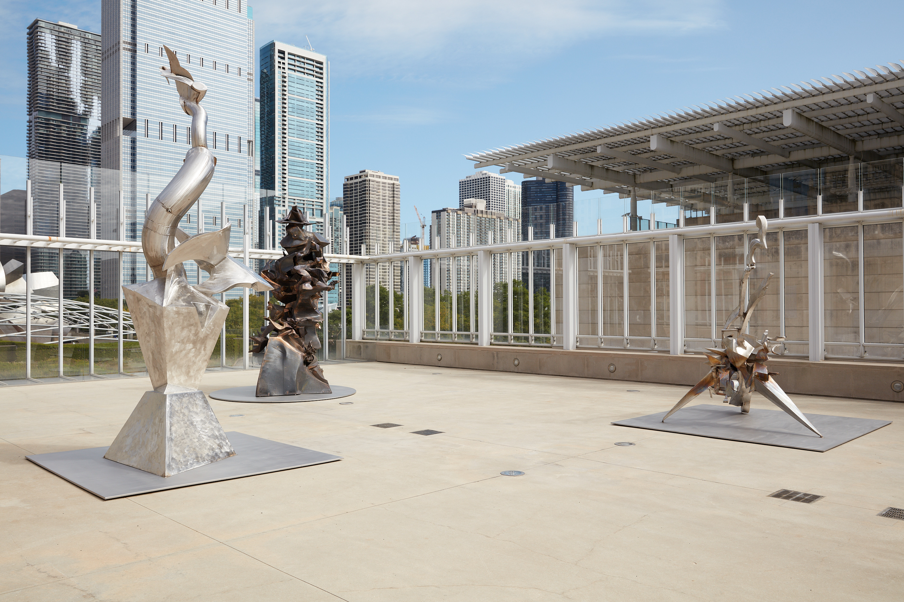 Three large twisting metal sculptures in various hues—silver, bronze, and golden—are installed on a concrete terrace. In the background skyscrapers and the flat roof of the adjacent building is seen..