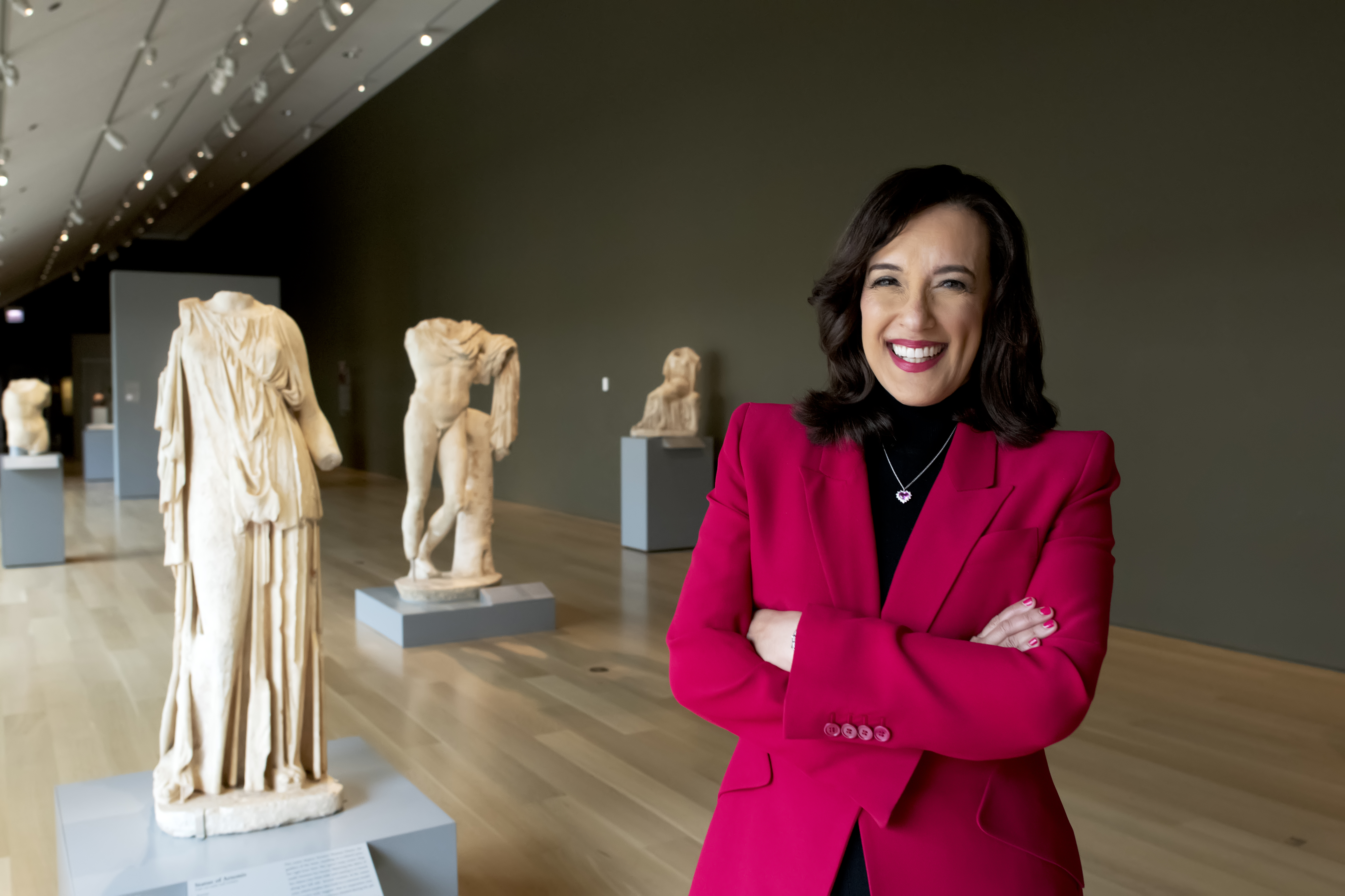Denise Gardner stands in a gallery space smiling with crossed arms, Classical sculptures visible behind her. She wears a bright-red suit jacket.