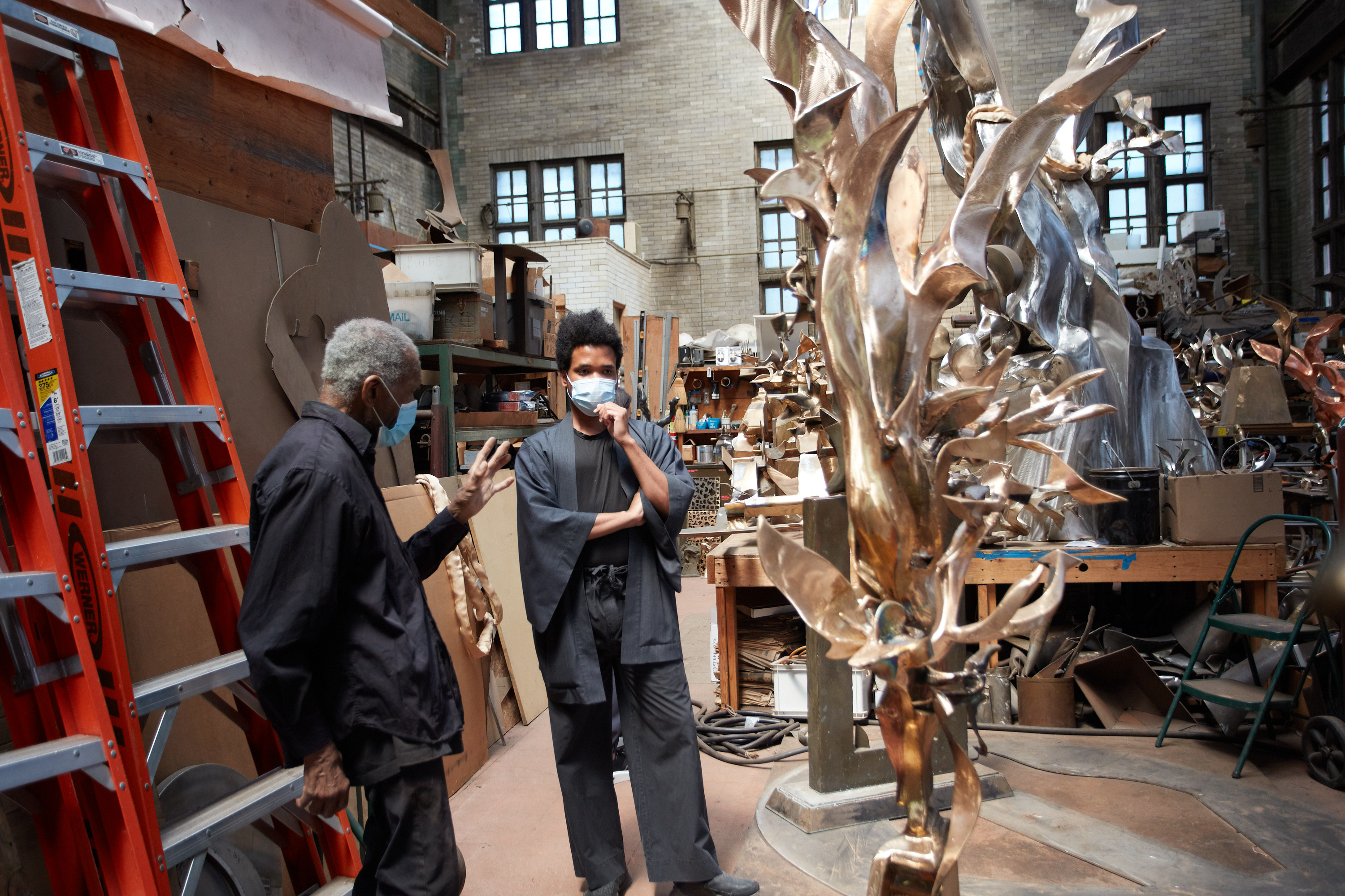 Sculptor Richard Hunt and curator Jordan Carter, both wearing face coverings, stand beside one of Hunt's bronze sculptures in his studio. Hunt leans against a red ladder.