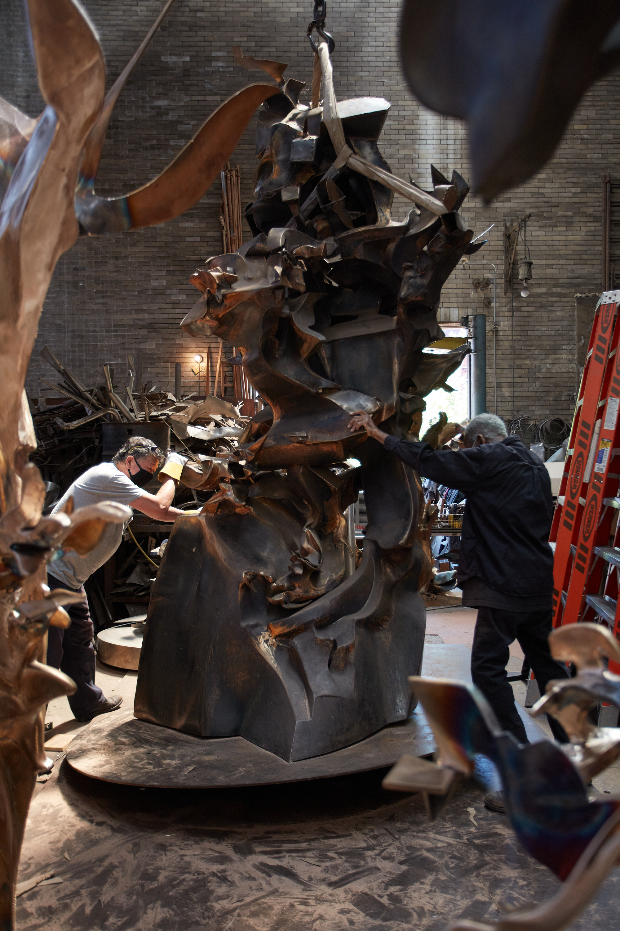 Two men push on either side of a tall, undulating bronze sculpture that appears to be lifted by a hook and cable from above.