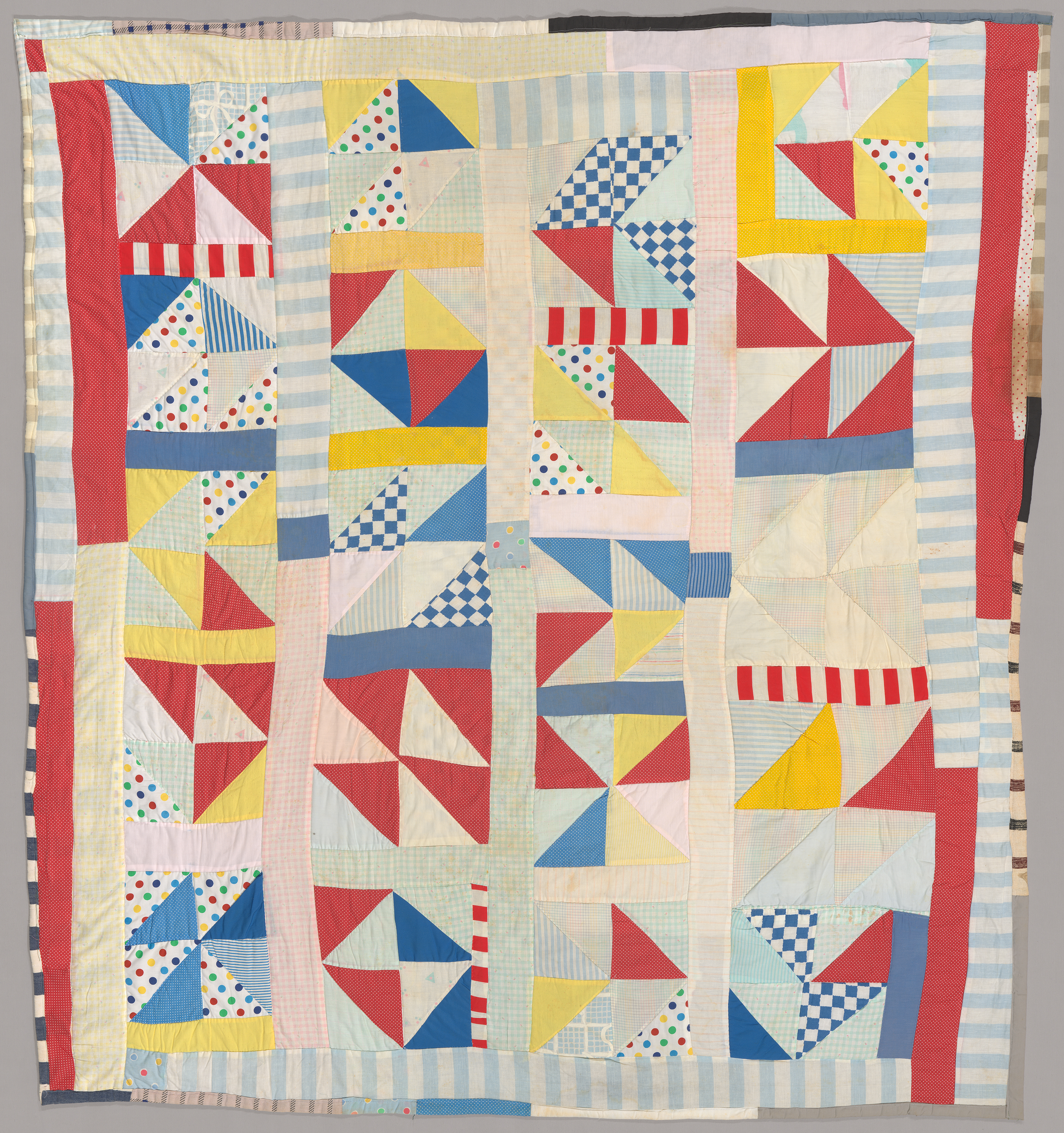 A quilt composed of solid red, yellow, blue, green, and light orange pieces as well as striped, checked, and polka-dotted fabric. Various triangular pieces create a dominant pinwheel-like pattern.