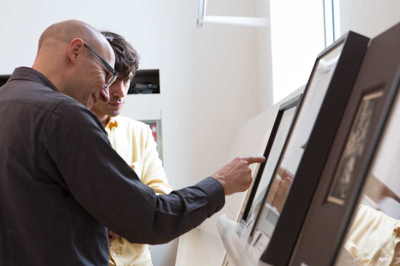 An older light-skinned man points to an artwork on the right of the image that we cannot see. A younger man is next to him in the background, also looking at the work.