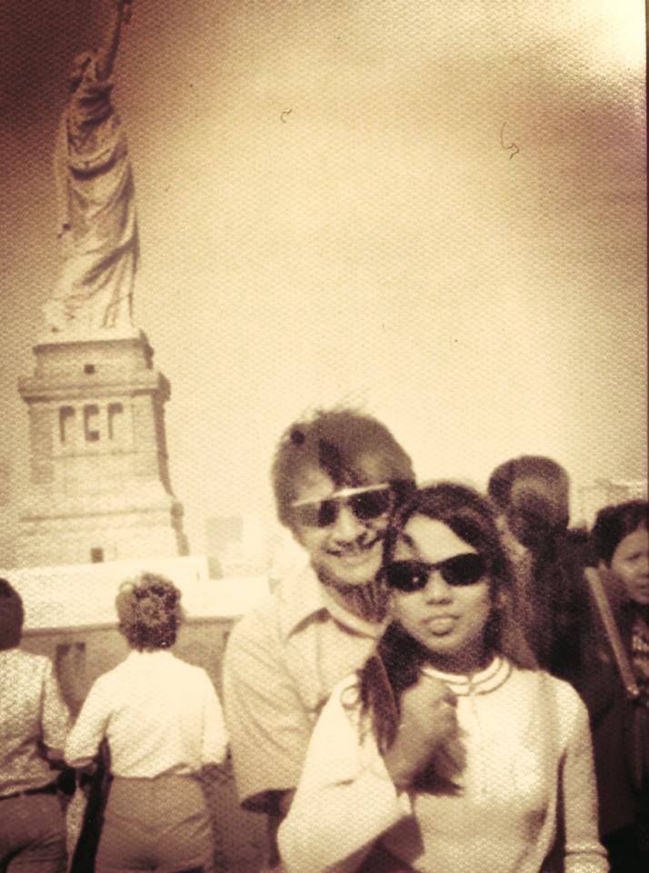 An Indian couple, both wearing sunglasses, pose together for a picture in front of the Statue of Liberty