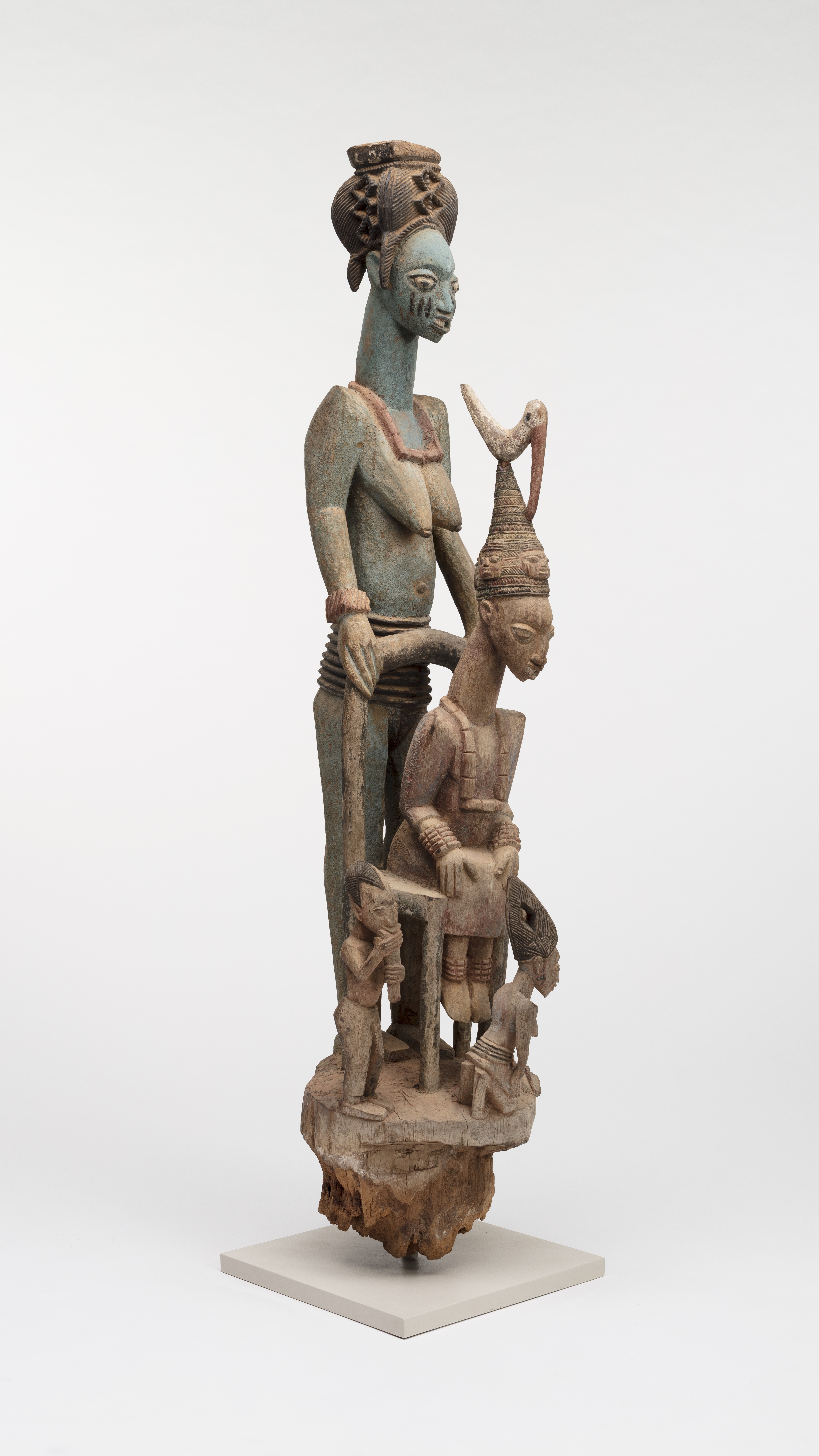 A carved and subtly colored wooden sculpture of a seated man wearing an elaborate headdress. A larger woman stands behind him and two much smaller figures at his feet.