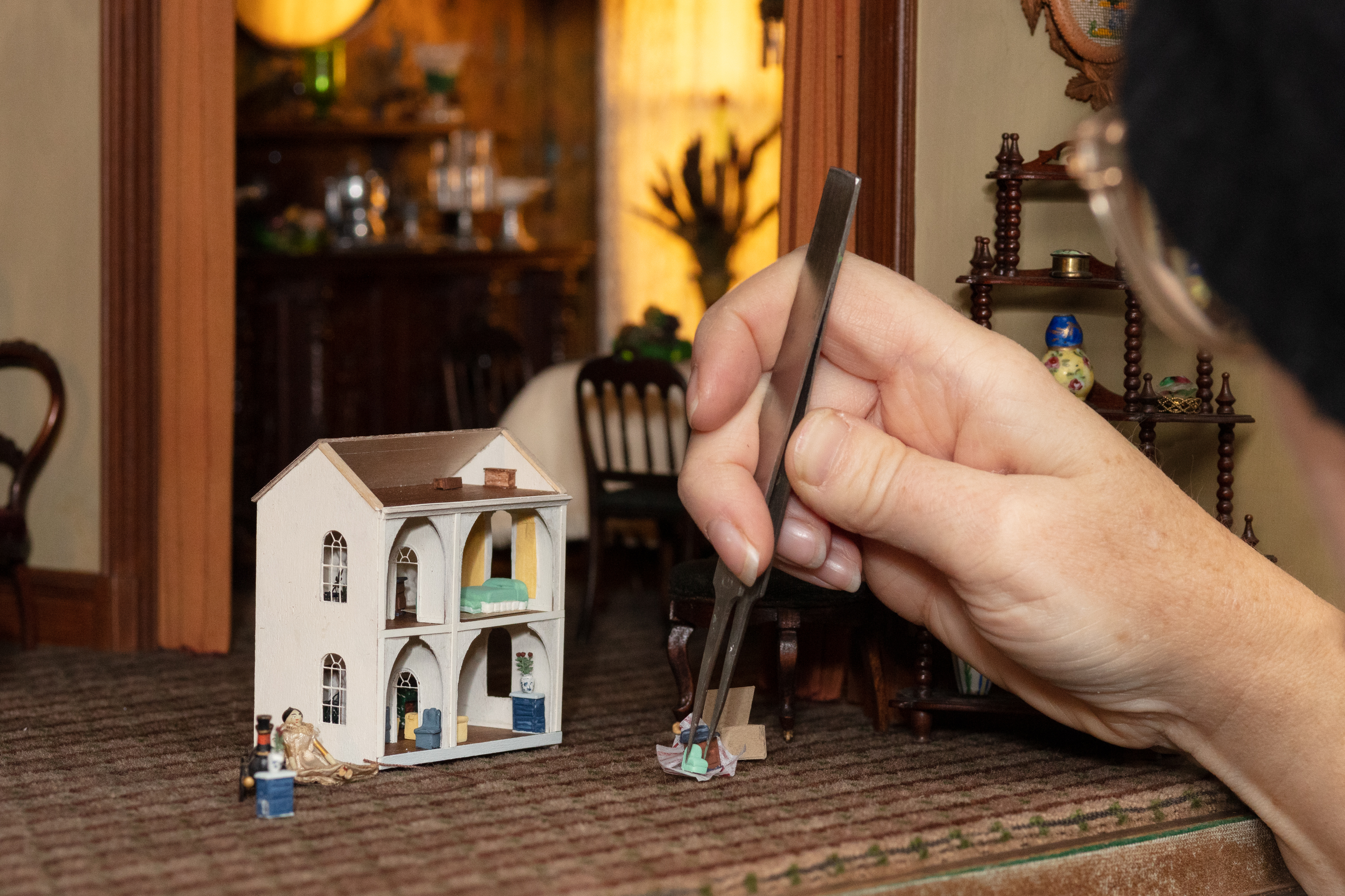 Photograph of hand holding tweezers and picking up a tiny blue chair in order to place into the tiny dollhouse that was made for the Thorne Miniature Rooms