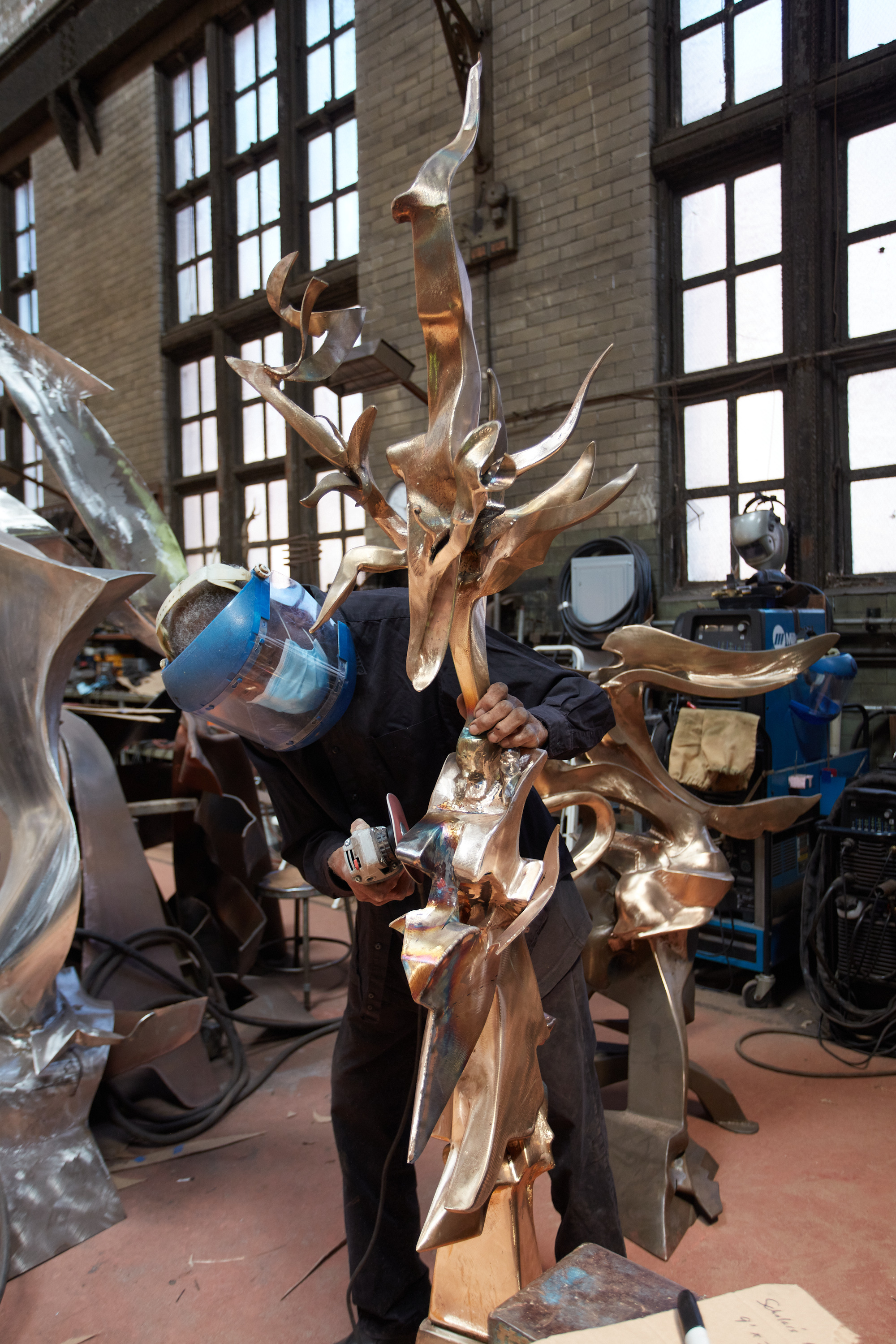 Sculptor Richard Hunt, wearing a face covering and face shield, bends over as he sands a bronze sculpture with numerous winding appendages.