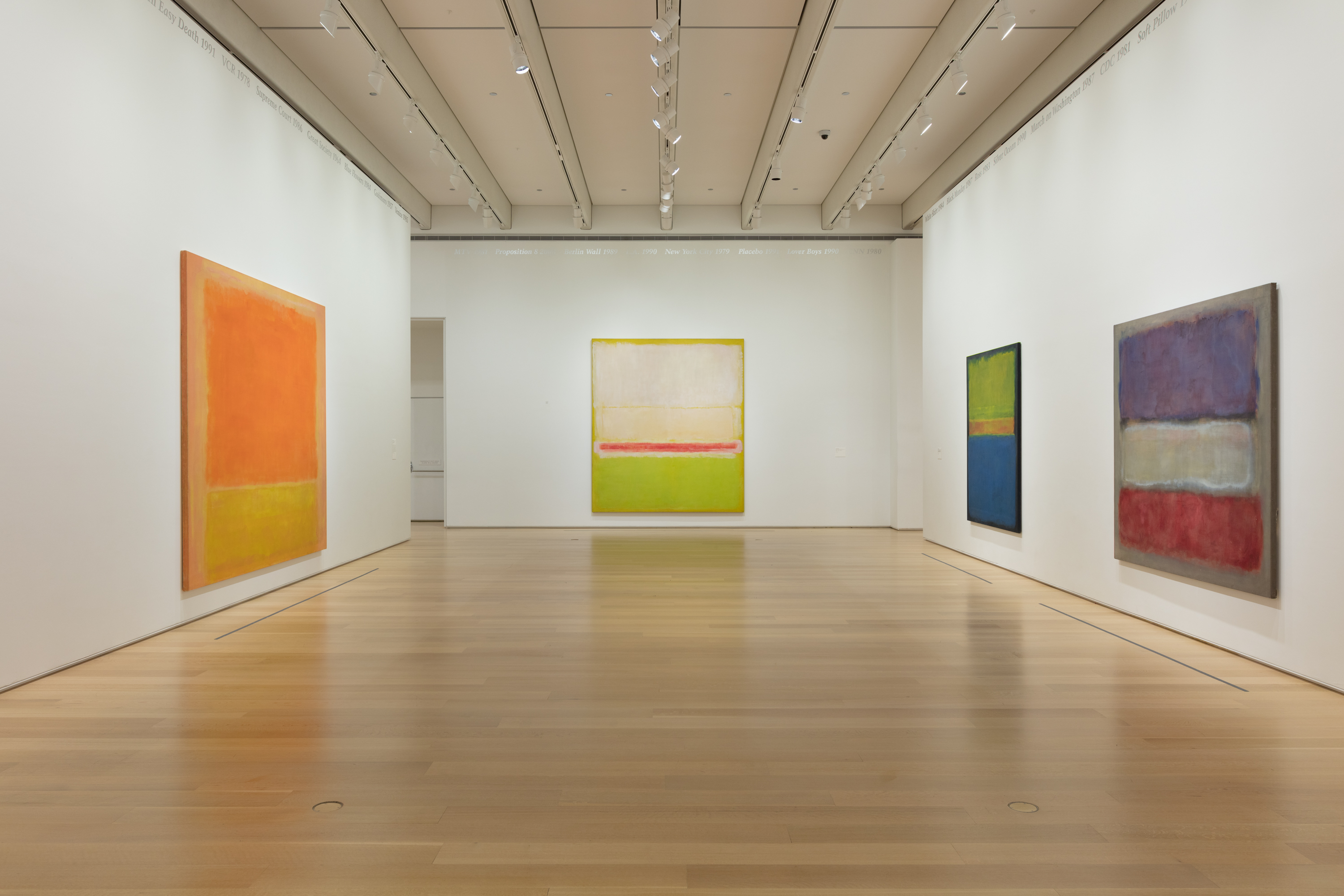 On three white walls of a gallery hang 4 large abstract painted canvases, each with horizontal bands of color that bleed together.