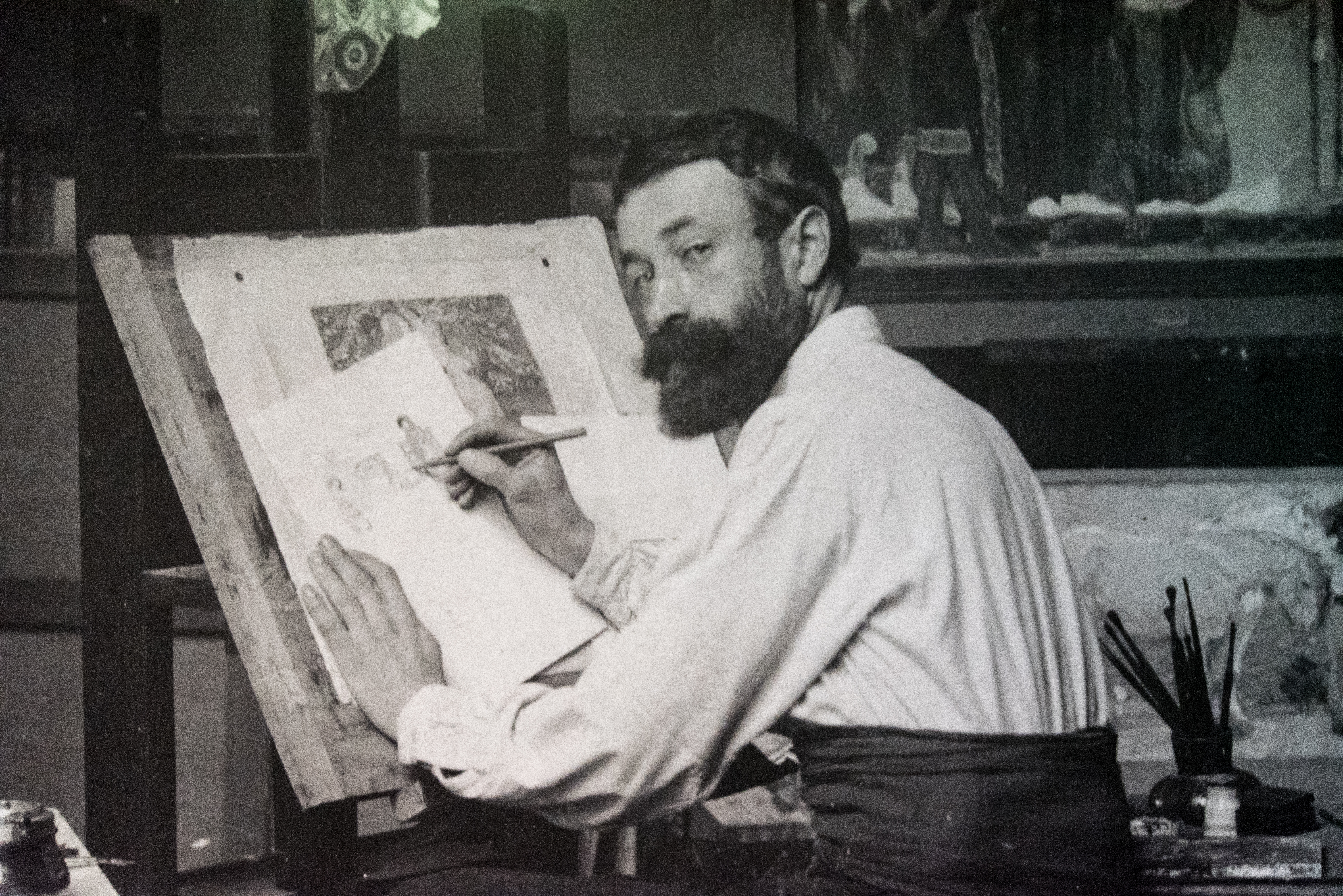 A male artist with short hair and neatly trimmed beard sits at a drawing board and turns towards the camera, pencil in hand.