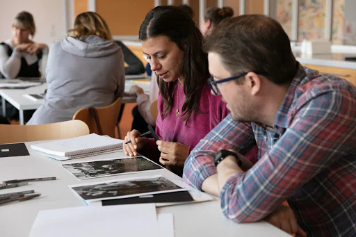 A woman and a man sit at a table and look at and discuss photographs that are laid on the table.