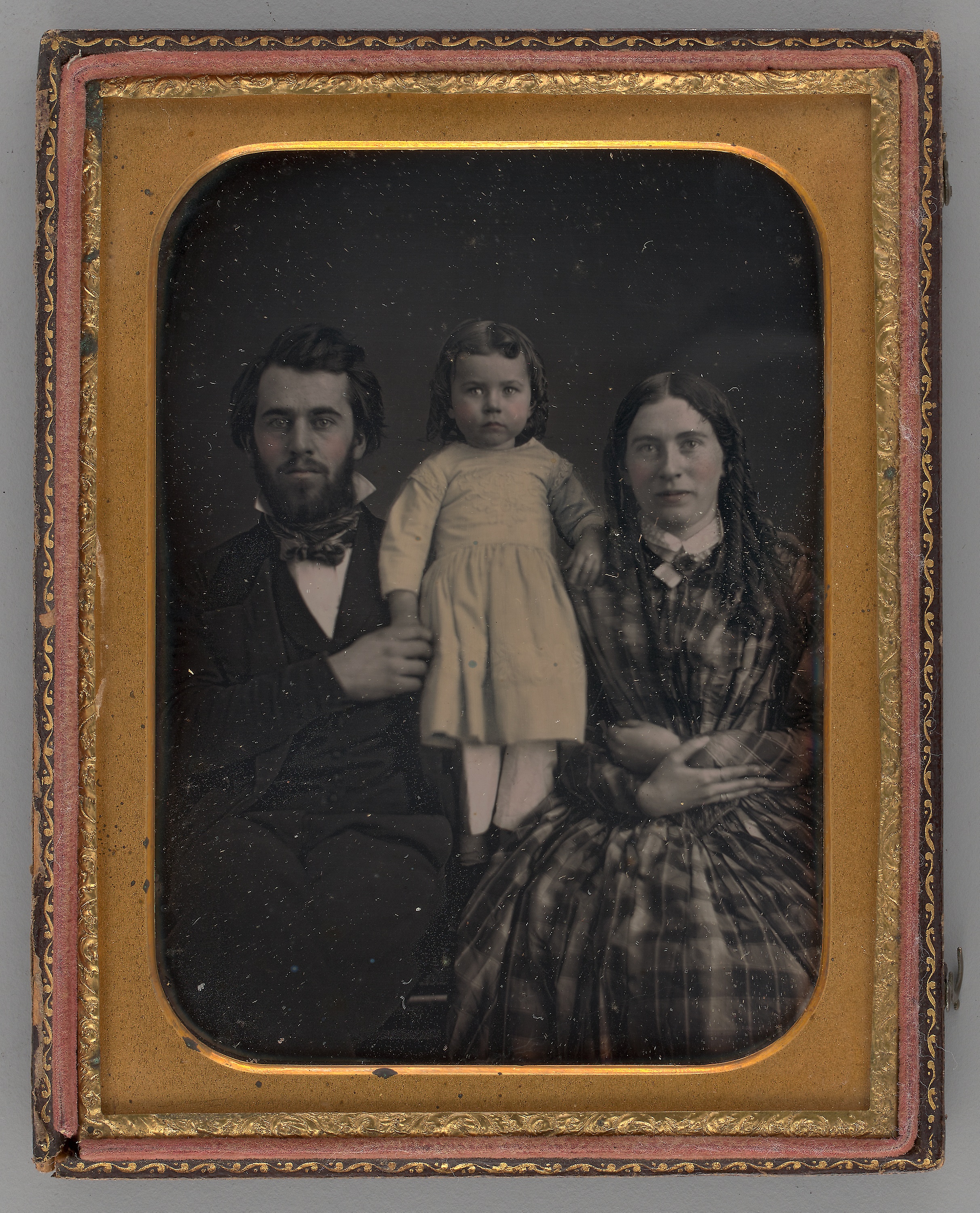 Black-and-white photograph of a family: a man on the left wears a suit, a child in the middle wears a white dress, and a woman on the right has long hair and wears a dress. They look directly into the camera with solemn expressions.