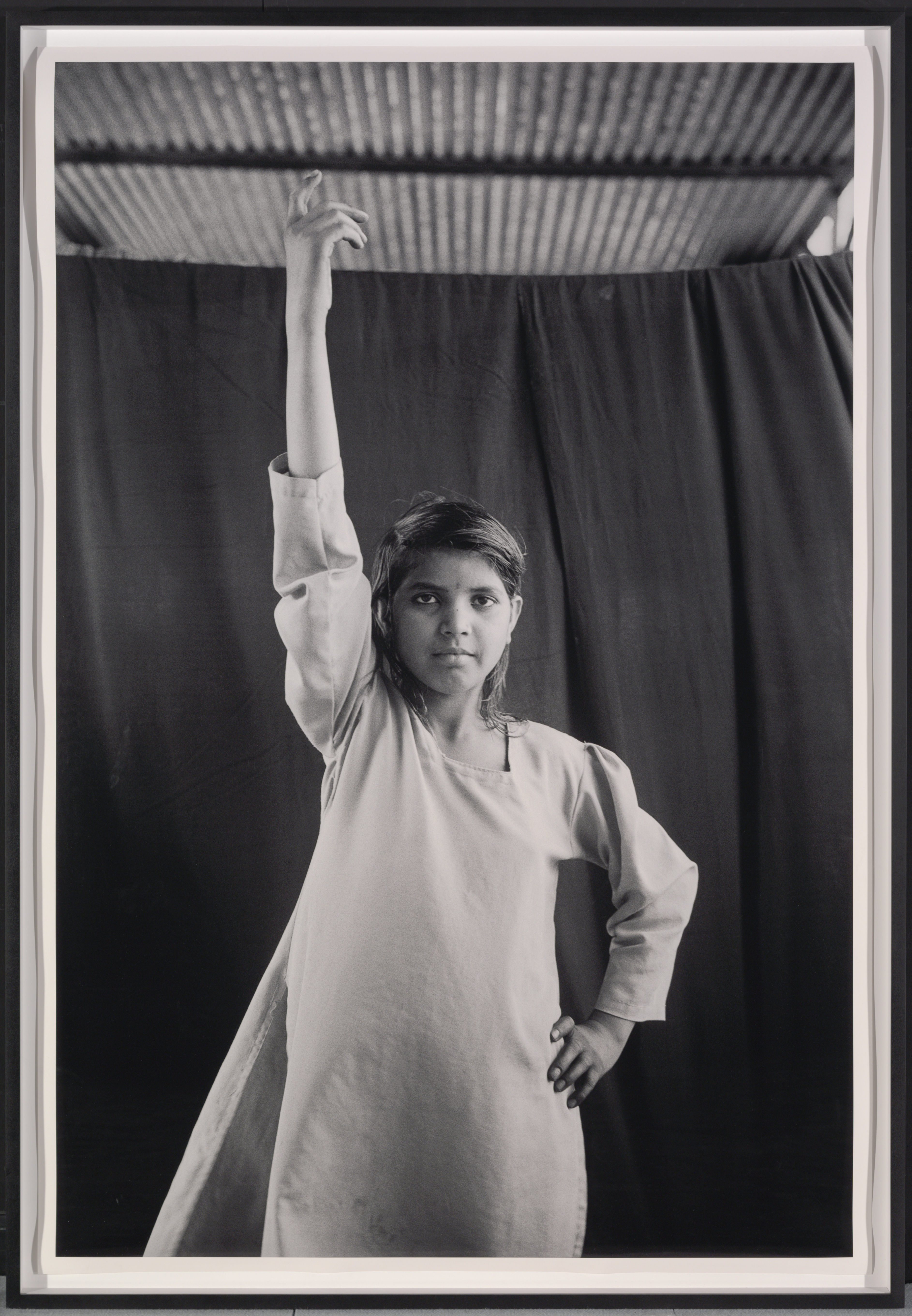 A black-and-white photo of young medium dark–skinned girl stands confidently facing the camera, her right arm raised and her left on hip. arm