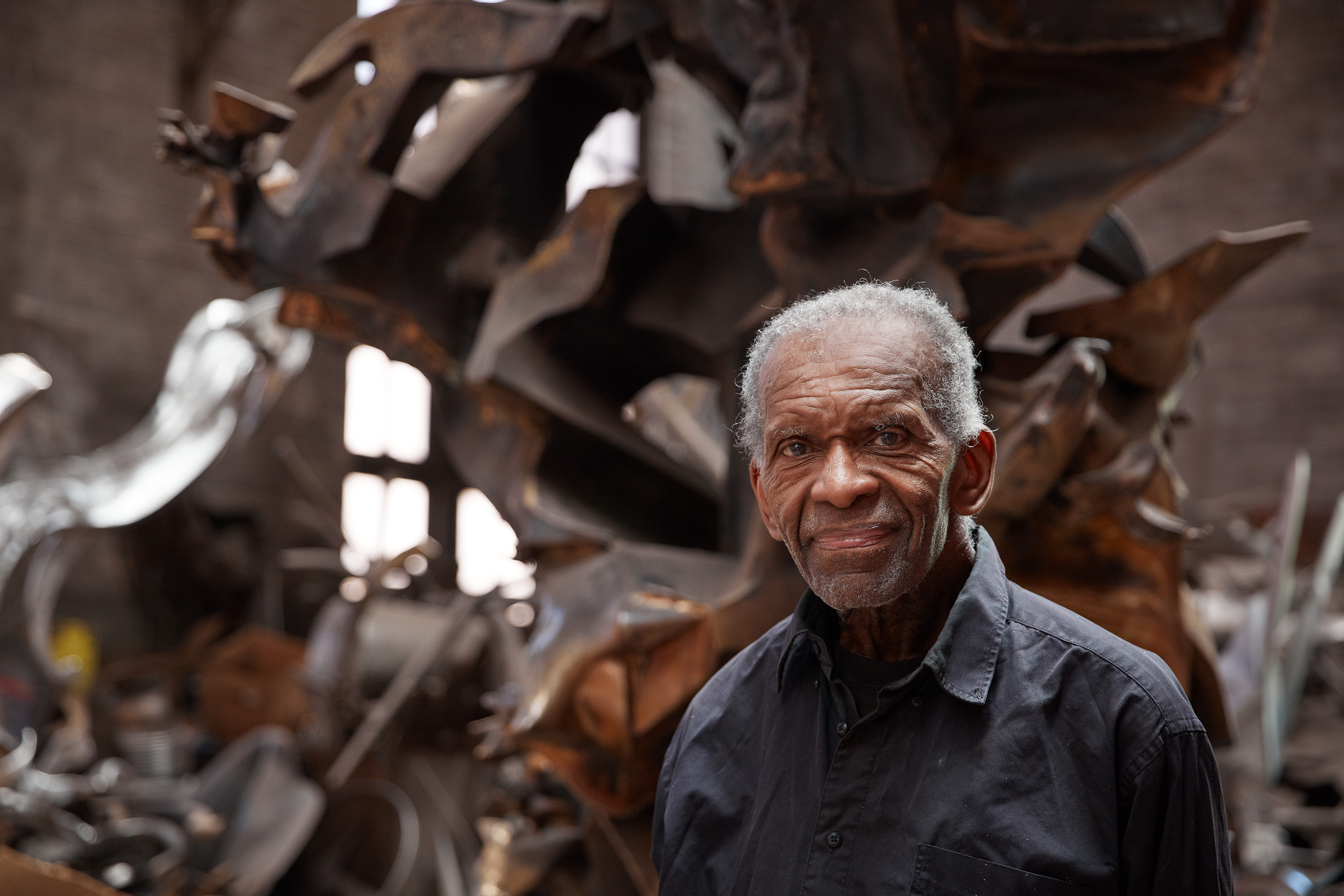 A photograph of artist Richard Hunt in his studio in front of his work, large-scale sculptures that twist and turn their way skyward.