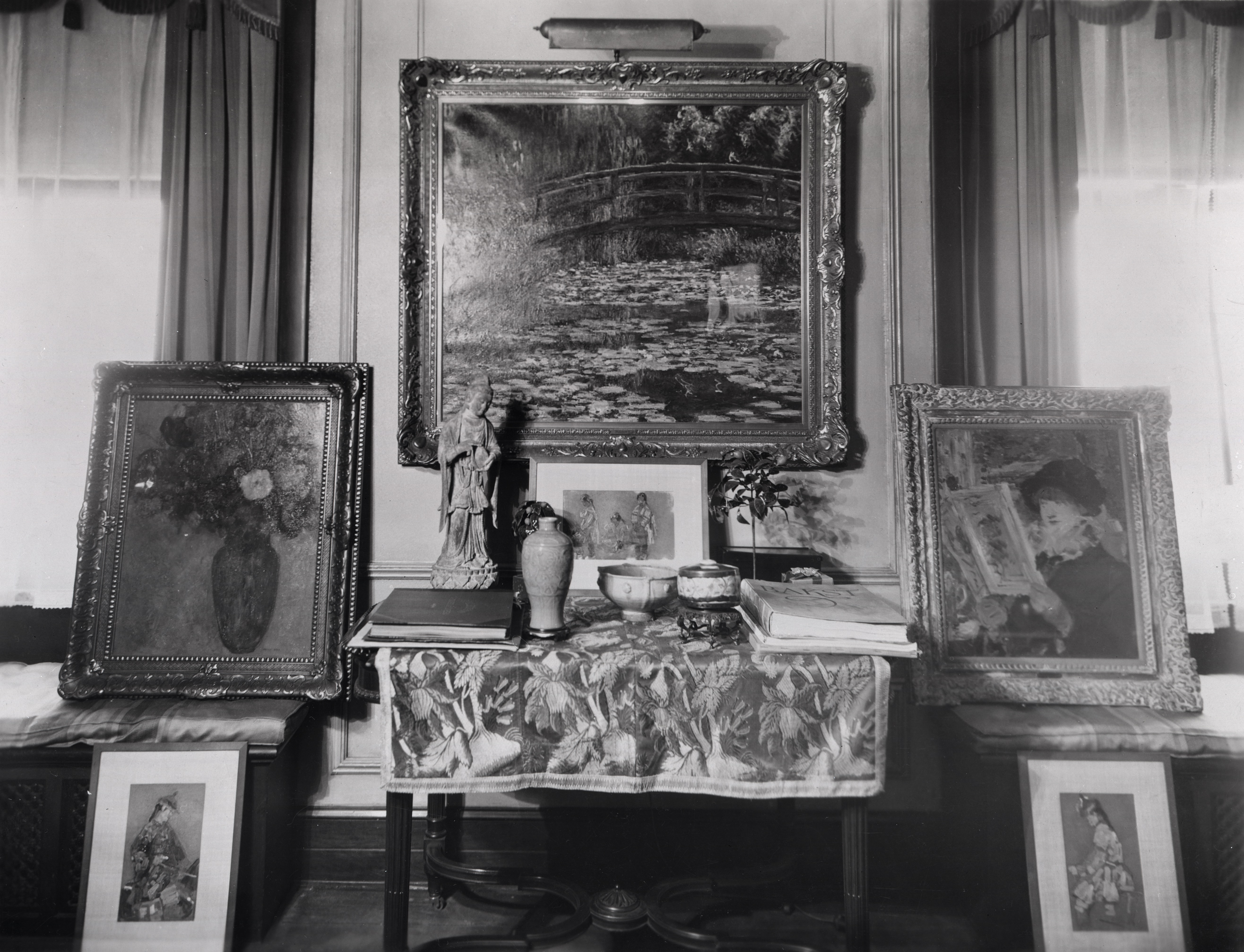 Black-and-white photograph of six paintings arranged in a room, leaning against or on a small table and chairs. Monet's "Water Lily Pond" hangs between two windows at center.