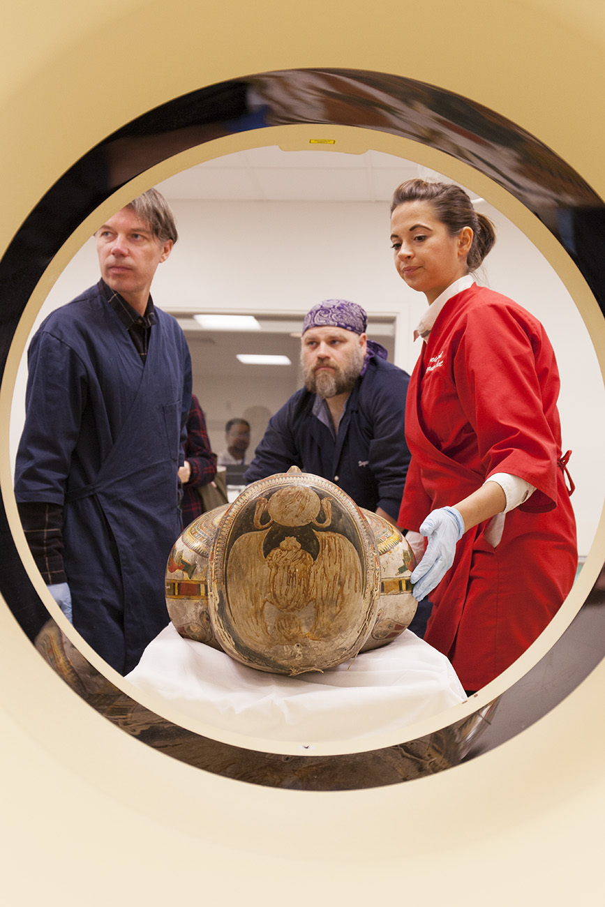 Photograph of three people putting an ancient Egyptian mummy case through a CT scanner. The mummy is laid flat. A man in a blue lab coat stands to the left, a man in a blue lab coat stands behind the mummy, and a woman in a red lab coat wearing blue gloves stands to the right.