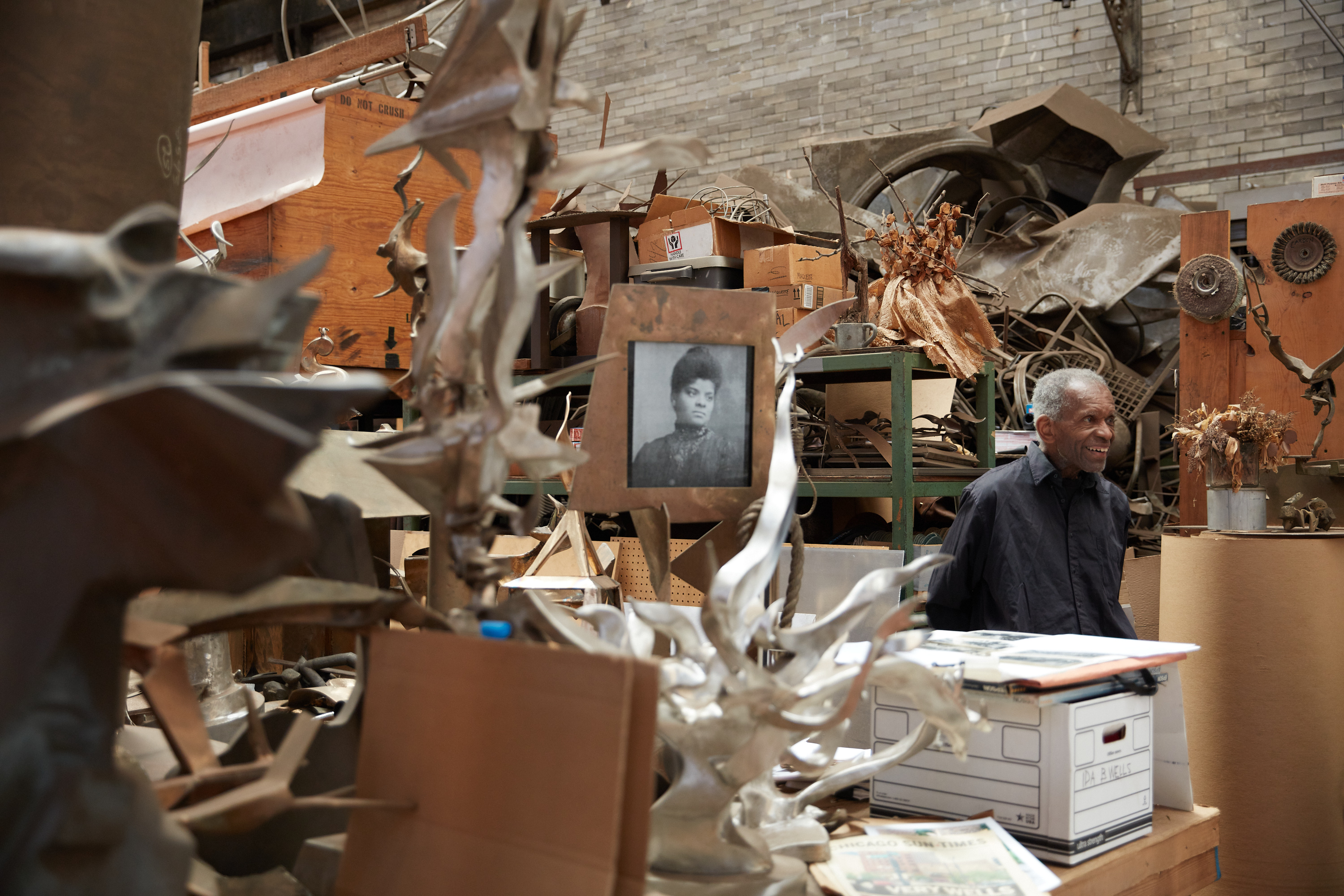 Sculptor Richard Hunt walks through his studio amid sculptures and piled boxes. A photograph of Ida B. Wells is seen prominently in the foreground.