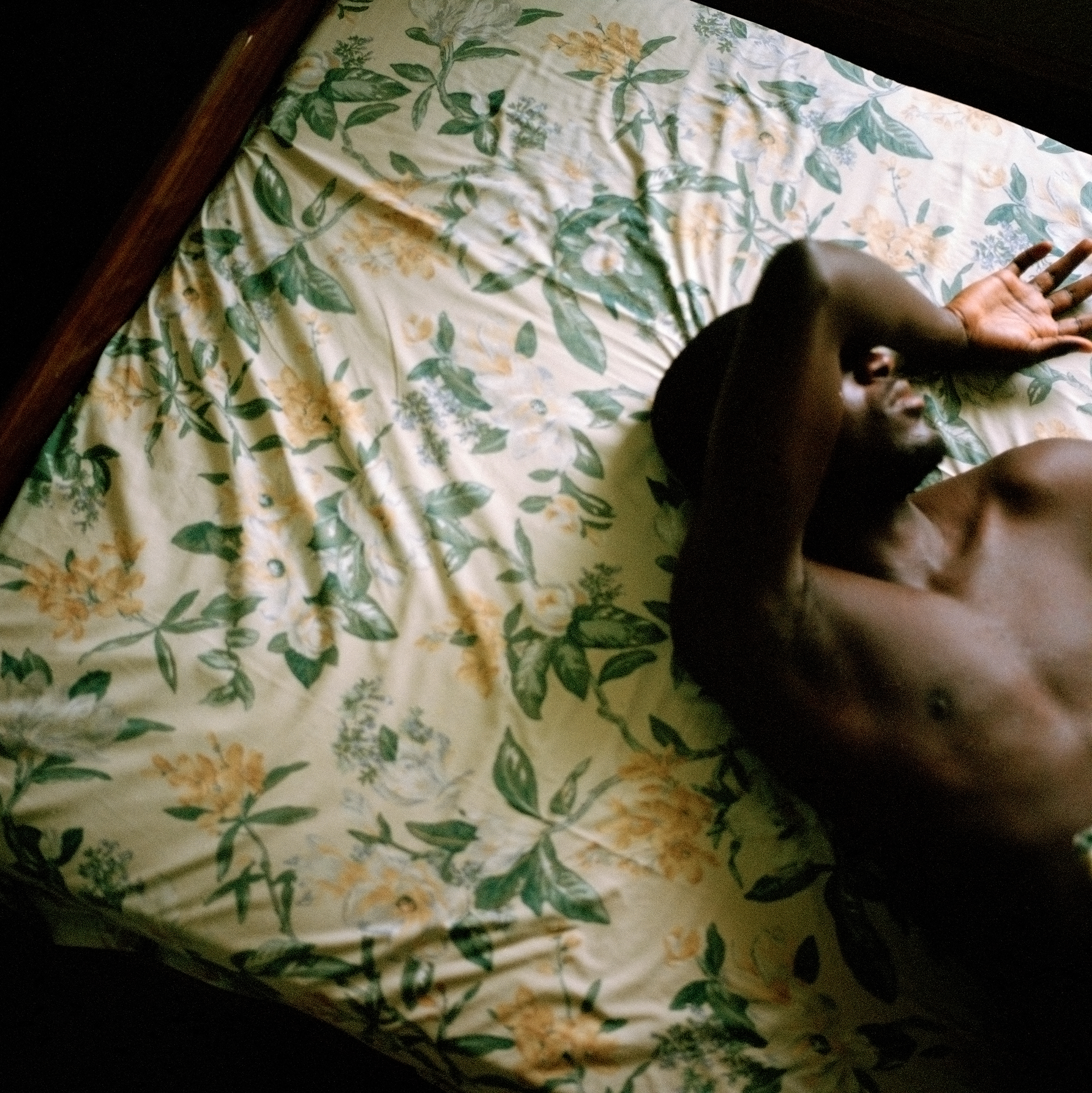 Photograph of a young man with dark brown skin lying on a bed with floral sheets in green and yellow. The man covers his eyes with his right arm, which is thrown over his face.
