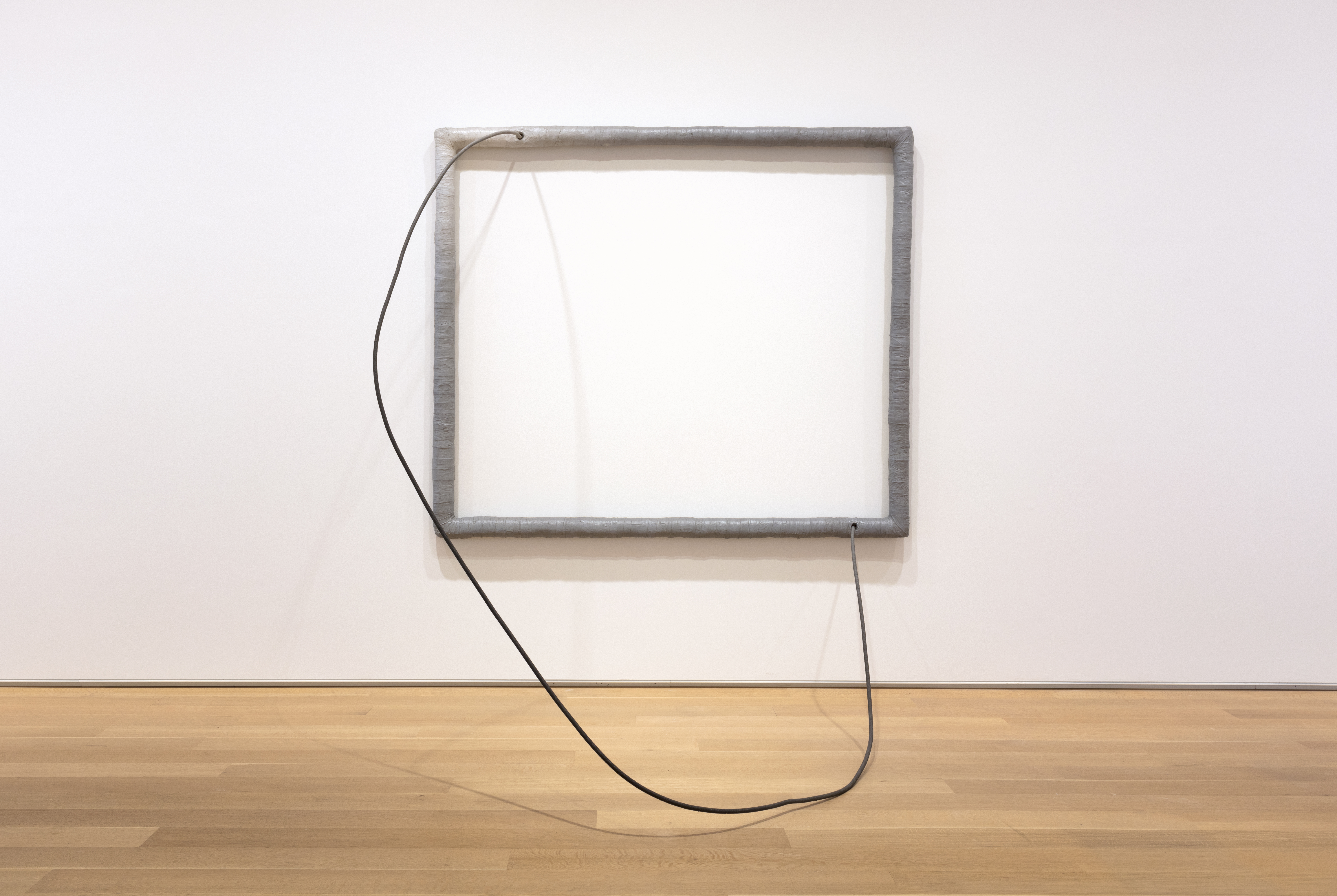 A photograph of a large, silver, metal frame hanging up on a stark white wall. A silver metal wire protrudes from the top left corner and ends near the right bottom corner of this empty frame.