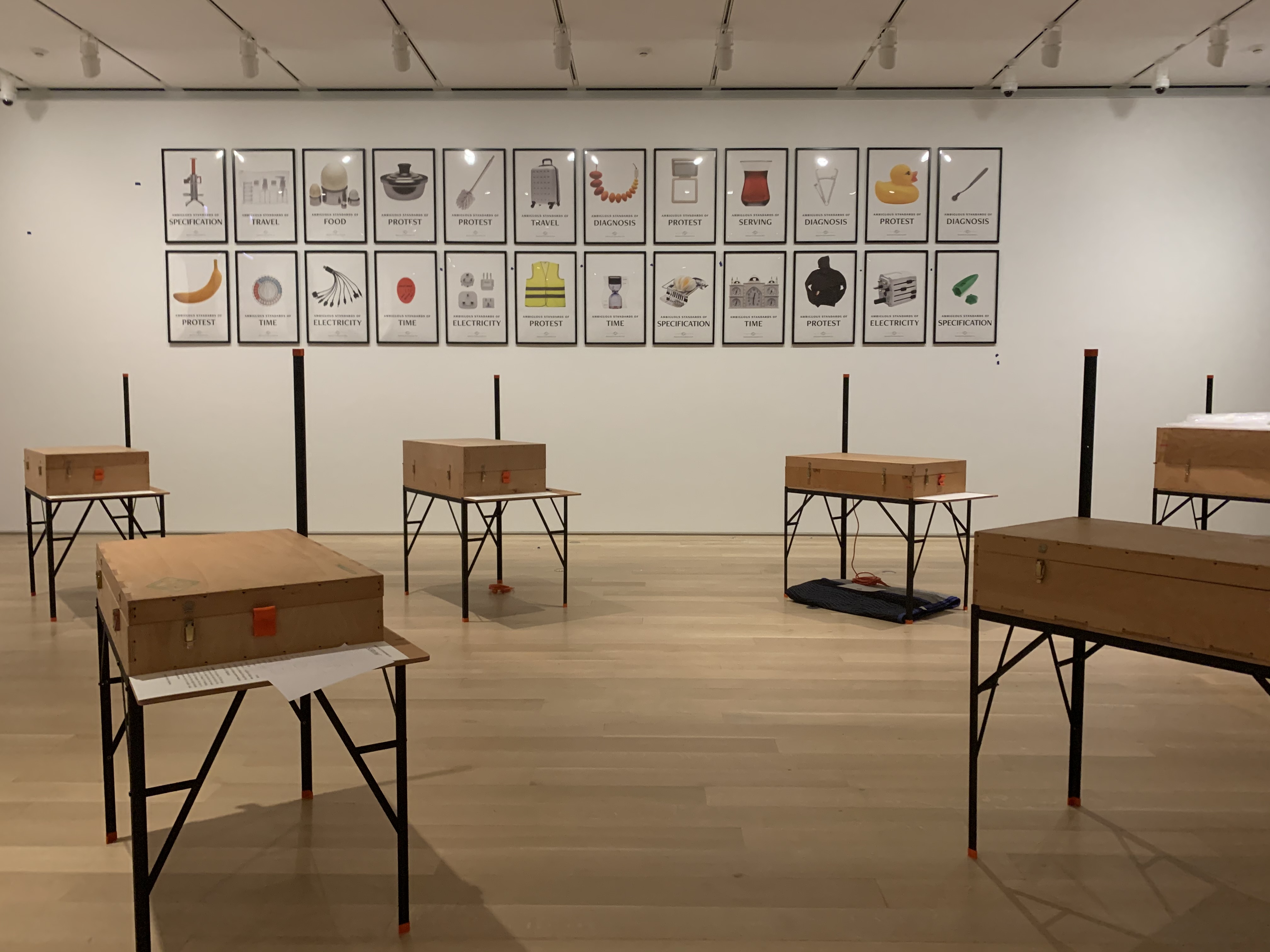 Numerous closed wooden boxes set on black metal frames fill a white gallery space. Two rows of posters populate the wall behind them.