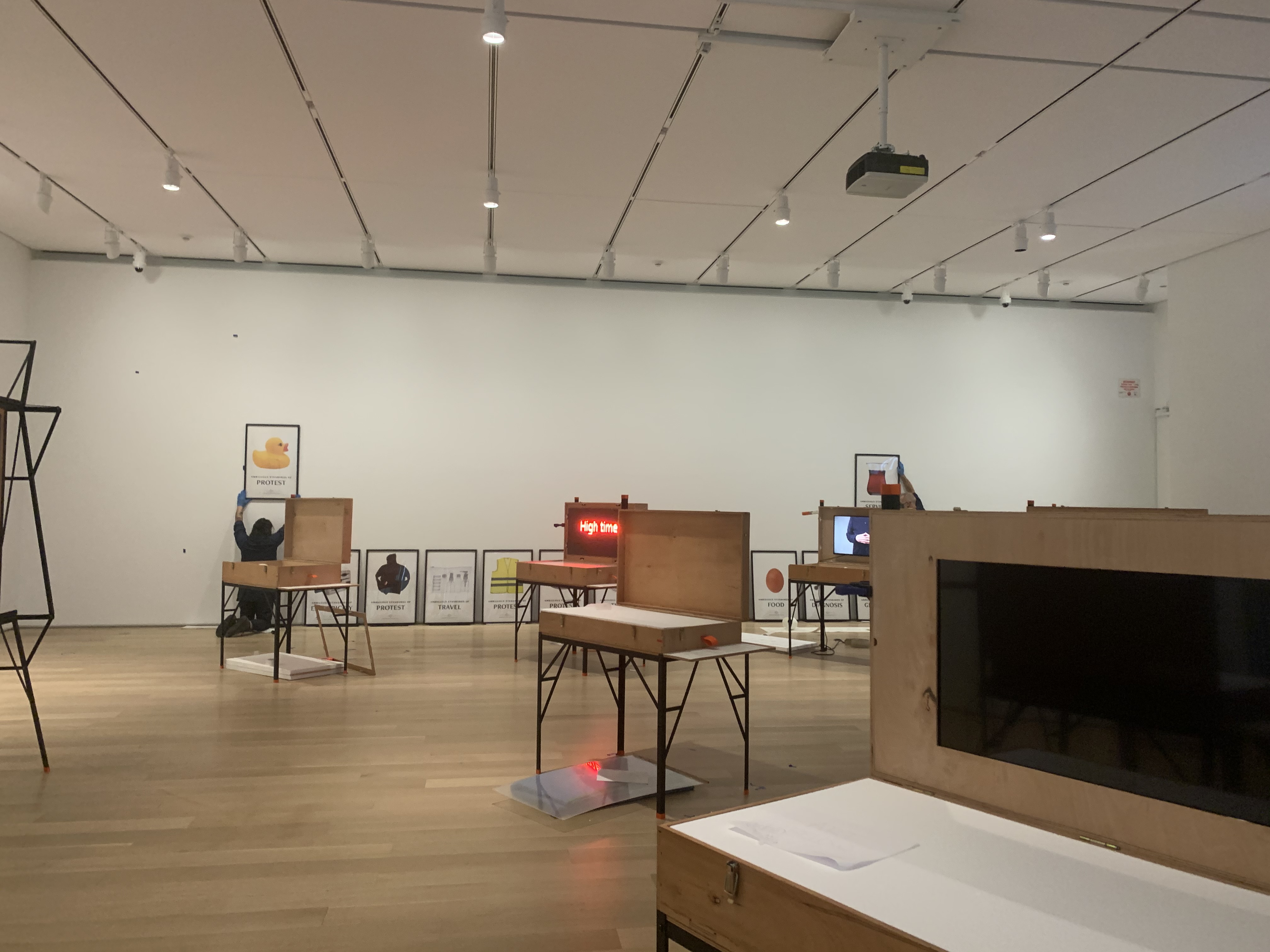Two figures hang posters on the far wall of a white gallery space filled with open wooden boxes set on metal frames.
