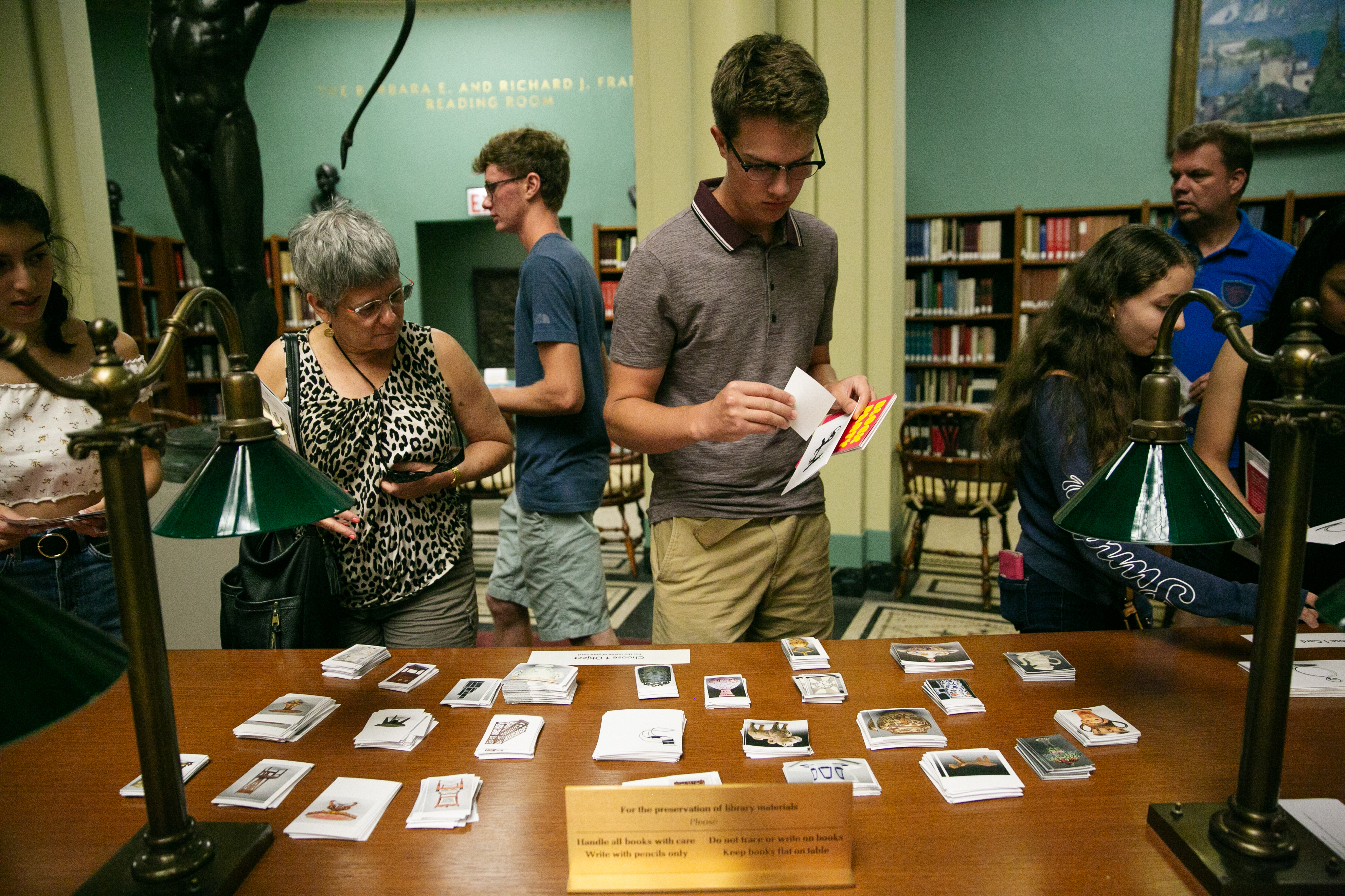 A photograph shows a wooden table top covered with small piles of postcards that various people are perusing and picking up. The space appears to be a library.