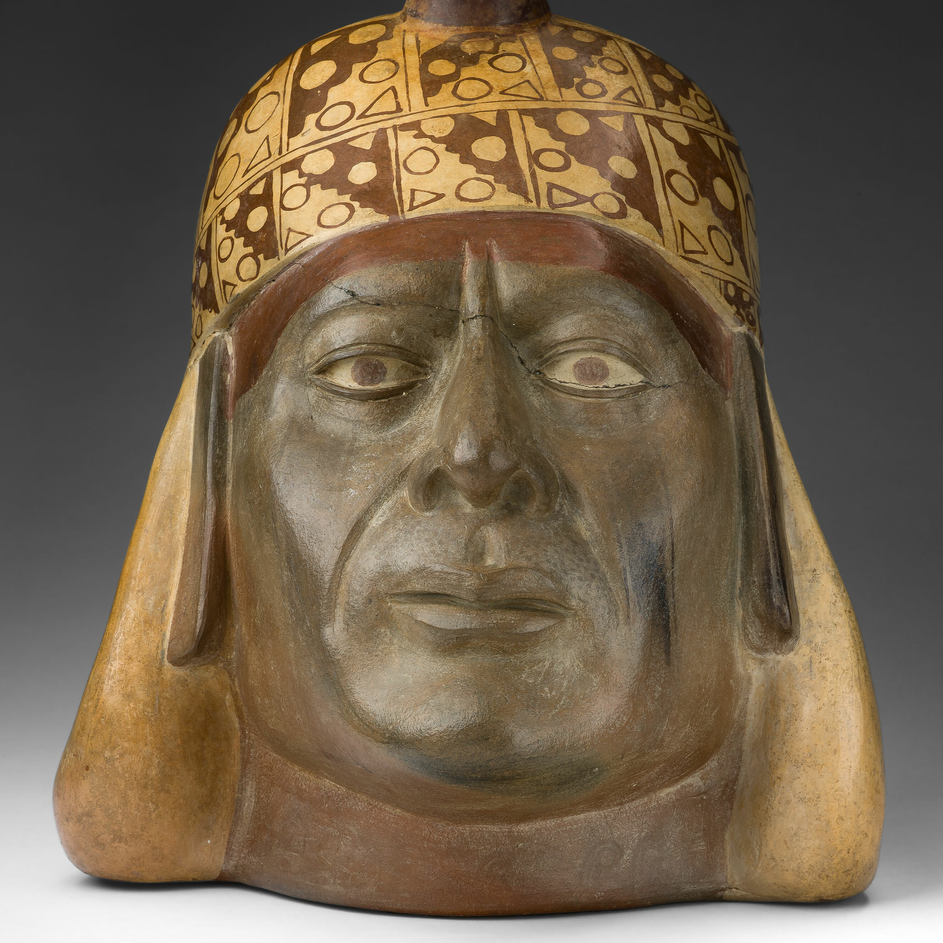 Sculpted head of a medium-skinned man in a beige headdress with geometric pattern and distinctive stirrup-shaped top piece.