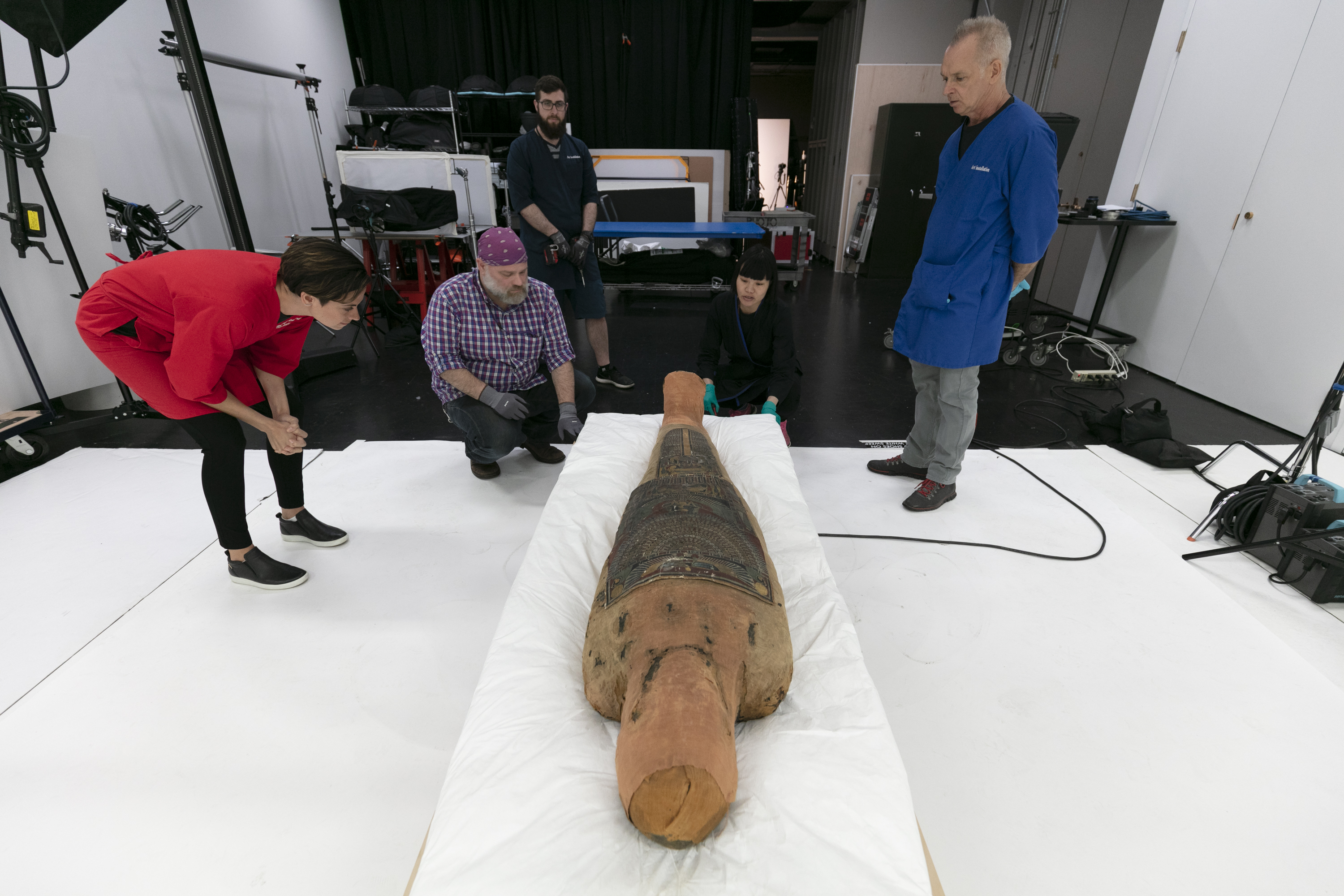 Photograph of a group of people examining an ancient Egyptian mummy case. The mummy case is flat, laid on a special pad. Five stand and kneel around the mummy case, examining it closely. The group includes three men and two women.
