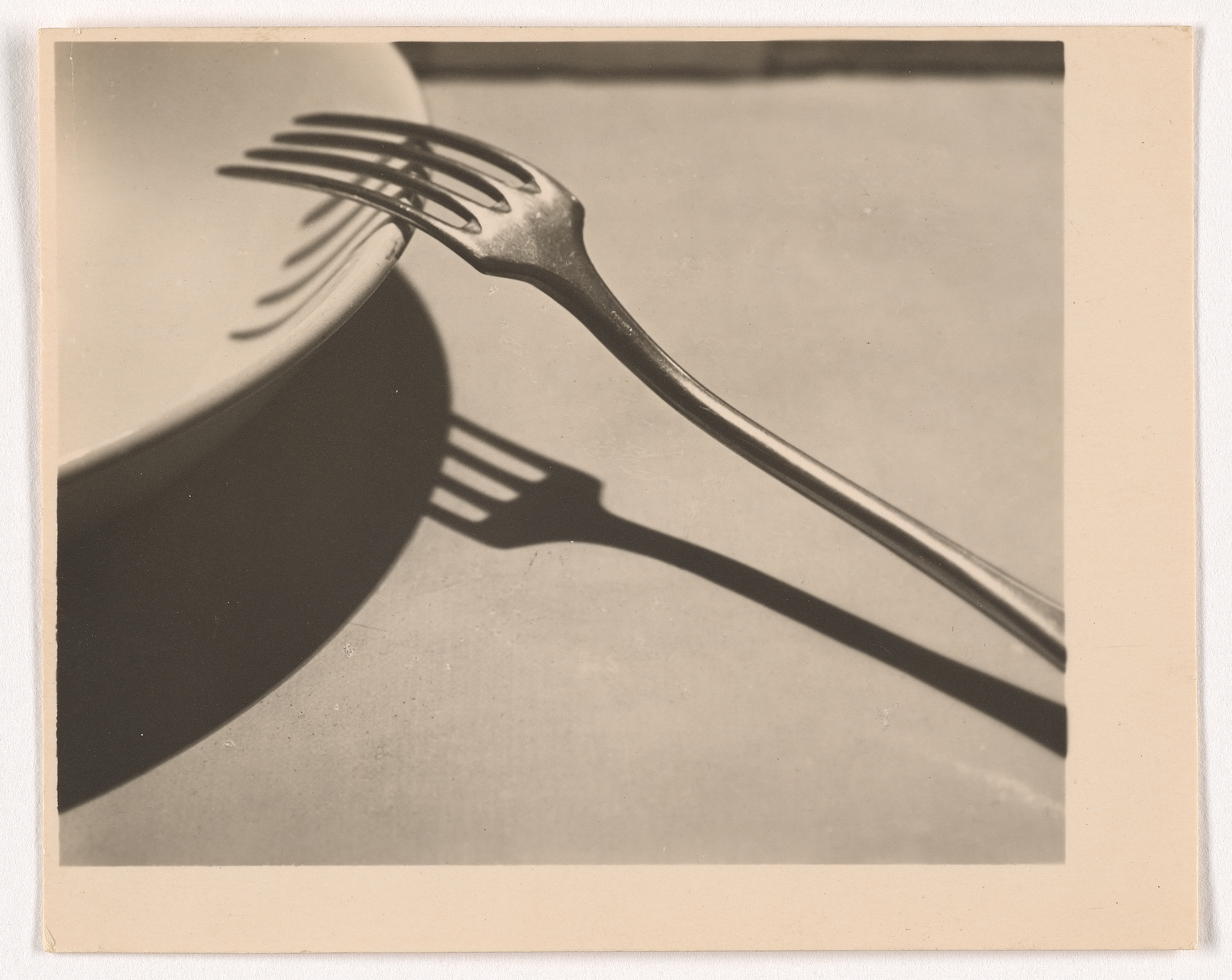 Black-and-white photograph of an upside-down fork, its prongs set on the lip of a pale, round dish. The fork casts a sharp shadow on the light surface below.